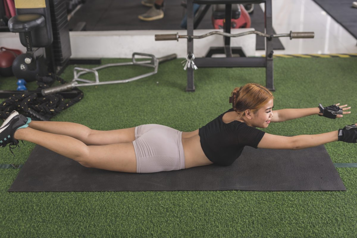 A Young Asian Lady Does Superman Back Extensions On A Black Mat. Isometric Exercise. Working Out And Training Lower Back Muscles At A Home Gym.