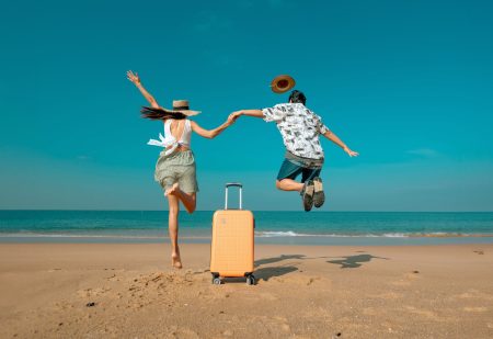 Last Minute Vakantie: man en vrouw maken een blije sprong op het strand, met tussen hen in een koffer