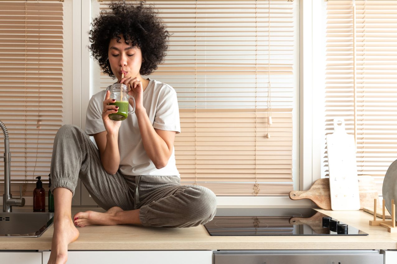 Mixed Race Black Latina Woman Drinking Healthy Green Juice Sitting On Kitchen Counter. Copy Space. Healthy Eating.