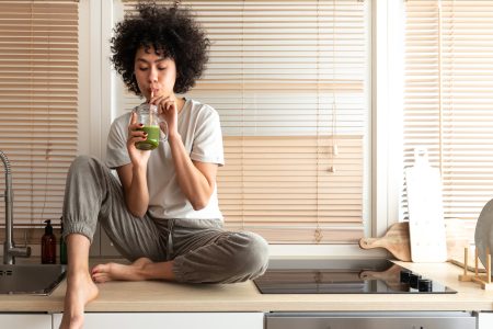 Mixed Race Black Latina Woman Drinking Healthy Green Juice Sitting On Kitchen Counter. Copy Space. Healthy Eating.