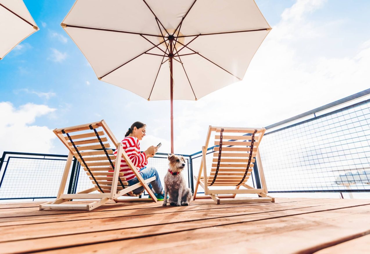 Vakantie Met Je Hond: Vrouw ontspant met haar telefoon op het terras met ligstoelen en parasols met haar hond