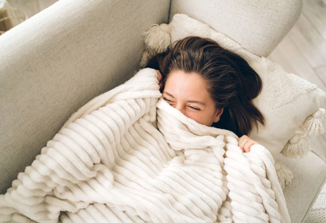 Warm Houden: Een jonge, lachende vrouw ligt op de bank onder een deken