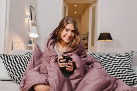 Pleased European Girl Drinking Tea On Sofa In Cold Day. Indoor Portrait Of Pretty Smiling Woman Sitting On Couch With Purple Blanket..