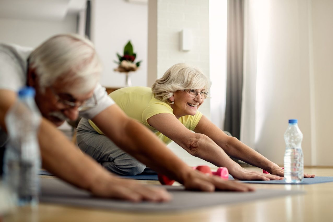 Happy Senior Woman And Her Husband Stretching On The Floor While Exercising At Home.