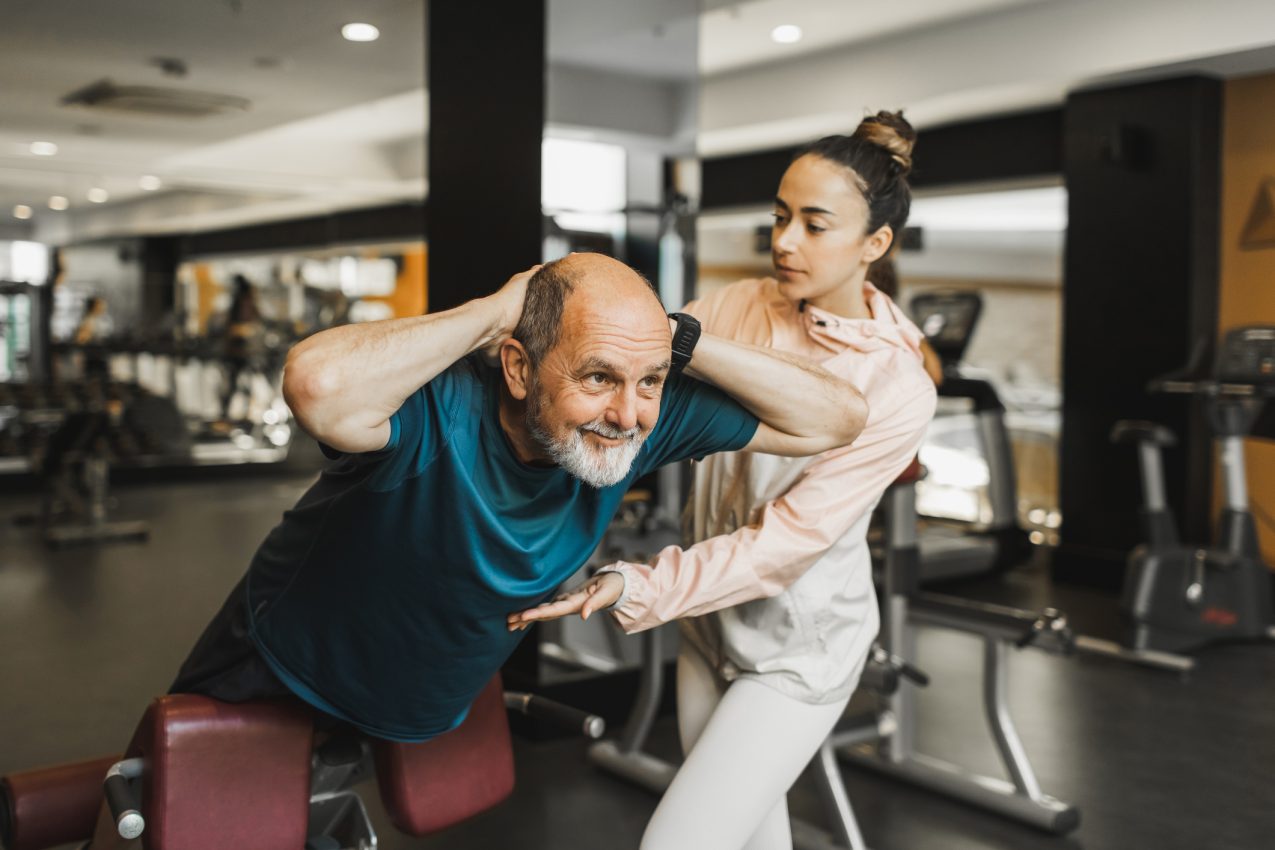 Old Man Doing Hyperextension Exercise For Strong Back And Young Coach Helping And Supporting Him