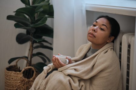 Central Heating Problems. Sad African American Woman Wrapped In Blanket Sitting Near Radiator At Home Holding With Cup Of Tea, Trying To Keep Warm In Cold Apartment